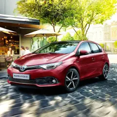 Czerwony Auris Hybrid, najbardziej ekonomiczne auto, sunie po brukowanej uliczce obok kawiarni.