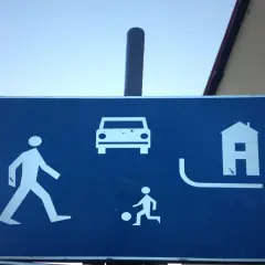 Znak informuje, że w strefie ruchu pieszy ma pierwszeństwo. Widać pieszego, dziecko grające w piłkę, samochód i dom.