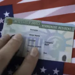 Dłoń trzyma zieloną kartę, dokument potwierdzający status stałego rezydenta USA, na tle amerykańskiej flagi. To dowód na to, co to zielona karta.