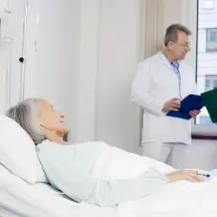 Starsza pacjentka w łóżku szpitalnym, lekarz i pielęgniarka omawiają jej stan, potencjalne czynniki jatrogenne.