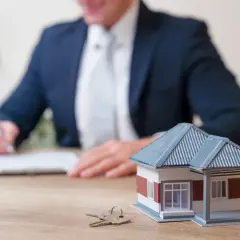 Model domu i klucze na biurku, a w tle mężczyzna w garniturze podpisuje dokumenty. Dowiedz się, jak ubezpieczyć mieszkanie.