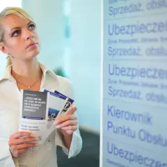Kobieta z ulotkami zastanawia się nad przedłużeniem OC przed terminem. Na tablicy widnieje napis "Ubezpieczenia".