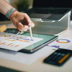 Ręka z rysikiem analizuje wykresy na tablecie. W tle laptop i dokumenty. Broker ubezpieczeniowy pracuje nad analizą danych.