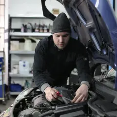 Mechanik w czapce z daszkiem pracuje przy silniku samochodu. W tle widać półki z płynami i beczki. To typowy warsztat, a nie miejsce na kosztorys.