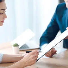 Kobieta podpisuje dokumenty dotyczące zmiany numeru rejestracyjnego a OC. Mężczyzna w garniturze podaje jej tablet.