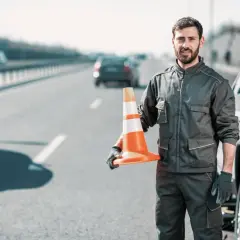 Pomoc drogowa na autostradzie. Mężczyzna w roboczym ubraniu trzyma pomarańczowy pachołek, gotowy do zabezpieczenia miejsca awarii auta.
