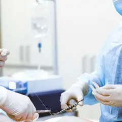 Chirurgiczne ręce w rękawiczkach szyją ranę. W tle kroplówka, ważna dla pacjenta, który może ubiegać się o odszkodowanie za operację.