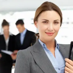 Uśmiechnięta bizneswoman z teczką, w tle współpracownicy omawiający dokumenty. Widać, że panuje między nimi umowa dorozumiana.