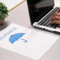 Przerwa w posiadaniu samochodu a zniżki na ubezpieczenie. Na biurku dokument "Insurance" z parasolką i osoba pisząca na laptopie.