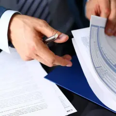 Ręce mężczyzny w garniturze przeglądają dokumenty, wśród których znajduje się formularz ubezpieczenia pośrednika nieruchomości.