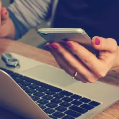 Kobieta sprawdza telefonem, czy nie ma nieprawidłowe dane w polisie OC. Na biurku leży laptop i pendrive.