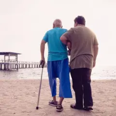 Mężczyzna z laską, wspierany przez drugiego, idzie po plaży. Może to symbolizować wsparcie w walce o odszkodowanie za stwardnienie rozsiane.