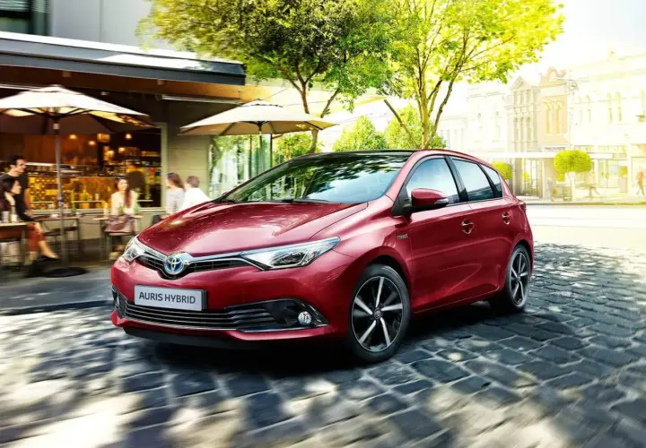 Czerwony Auris Hybrid, najbardziej ekonomiczne auto, sunie po brukowanej uliczce obok kawiarni.