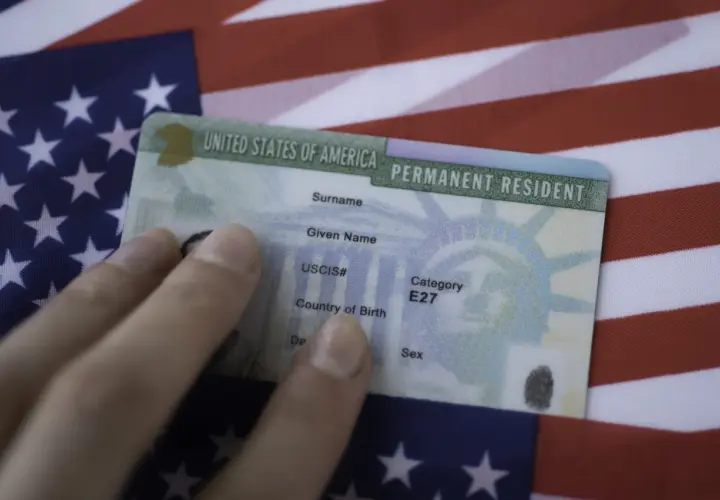 Dłoń trzyma zieloną kartę, dokument potwierdzający status stałego rezydenta USA, na tle amerykańskiej flagi. To dowód na to, co to zielona karta.