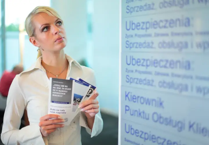 Kobieta z ulotkami zastanawia się nad przedłużeniem OC przed terminem. Na tablicy widnieje napis "Ubezpieczenia".