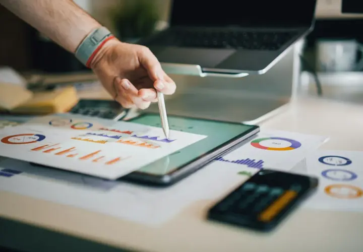 Ręka z rysikiem analizuje wykresy na tablecie. W tle laptop i dokumenty. Broker ubezpieczeniowy pracuje nad analizą danych.