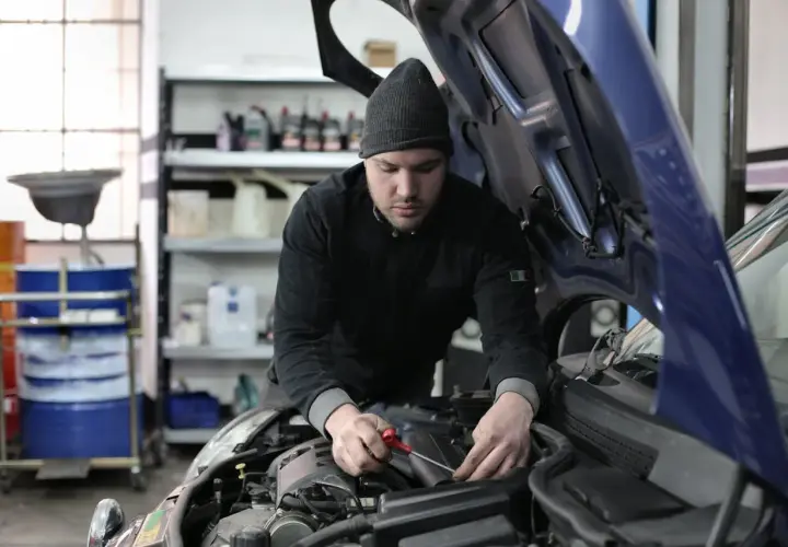 Mechanik w czapce z daszkiem pracuje przy silniku samochodu. W tle widać półki z płynami i beczki. To typowy warsztat, a nie miejsce na kosztorys.