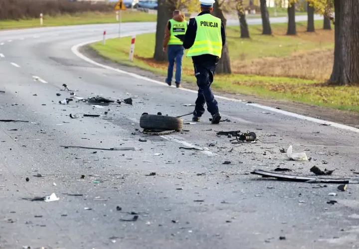 Na drodze po wypadku leżą szczątki pojazdów i opona. Policjanci w kamizelkach odblaskowych badają miejsce zdarzenia, być może szukając śladów ucieczki z miejsca wypadku.