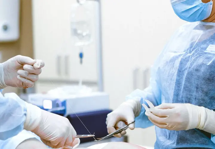 Chirurgiczne ręce w rękawiczkach szyją ranę. W tle kroplówka, ważna dla pacjenta, który może ubiegać się o odszkodowanie za operację.
