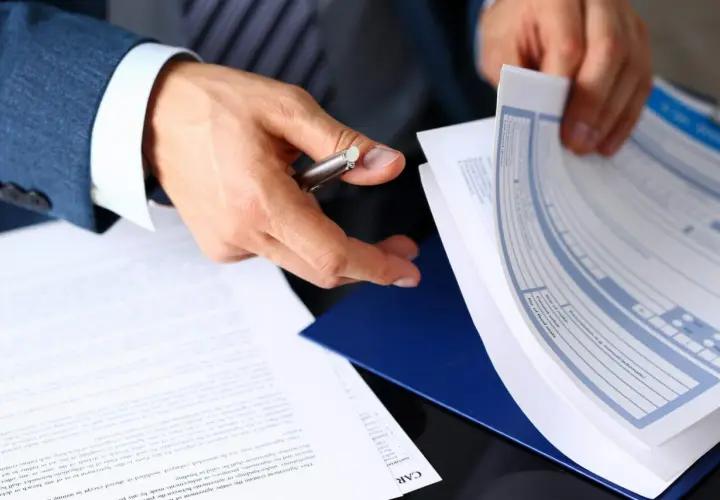 Ręce mężczyzny w garniturze przeglądają dokumenty, wśród których znajduje się formularz ubezpieczenia pośrednika nieruchomości.