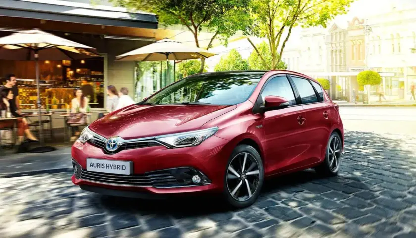 Czerwony Auris Hybrid, najbardziej ekonomiczne auto, sunie po brukowanej uliczce obok kawiarni.