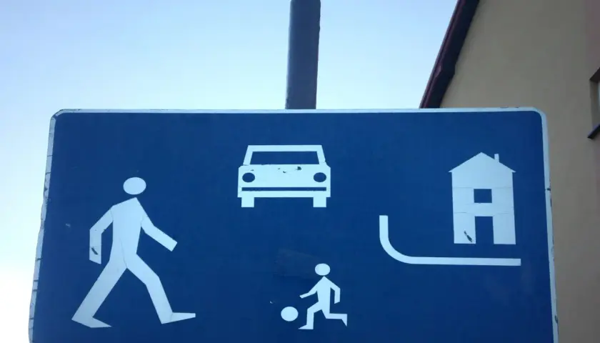 Znak informuje, że w strefie ruchu pieszy ma pierwszeństwo. Widać pieszego, dziecko grające w piłkę, samochód i dom.