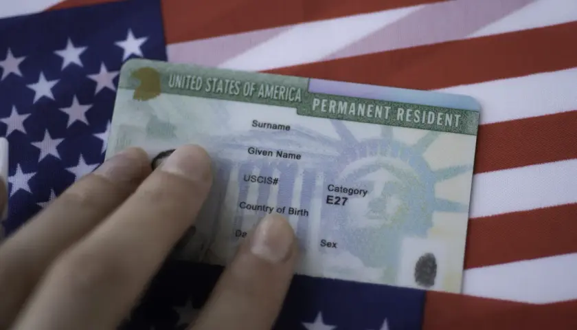 Dłoń trzyma zieloną kartę, dokument potwierdzający status stałego rezydenta USA, na tle amerykańskiej flagi. To dowód na to, co to zielona karta.