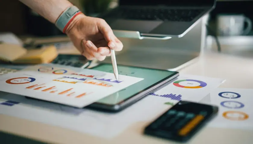 Ręka z rysikiem analizuje wykresy na tablecie. W tle laptop i dokumenty. Broker ubezpieczeniowy pracuje nad analizą danych.