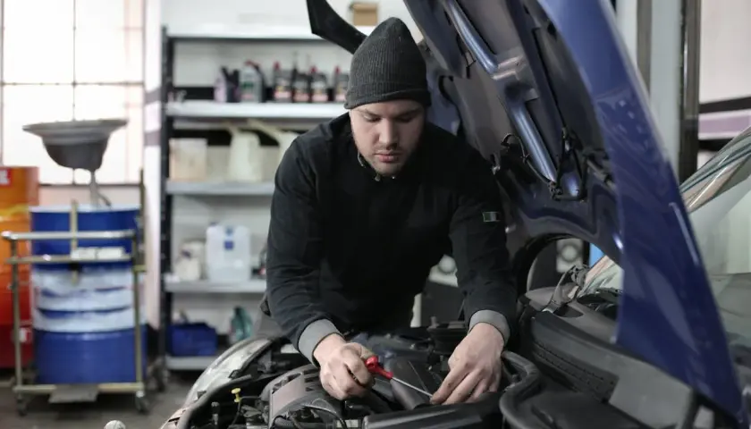 Mechanik w czapce z daszkiem pracuje przy silniku samochodu. W tle widać półki z płynami i beczki. To typowy warsztat, a nie miejsce na kosztorys.