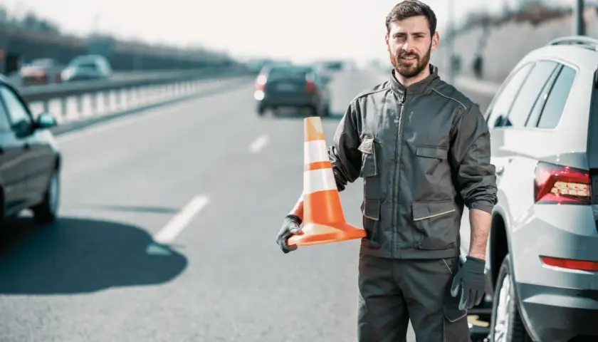 Pomoc drogowa na autostradzie. Mężczyzna w roboczym ubraniu trzyma pomarańczowy pachołek, gotowy do zabezpieczenia miejsca awarii auta.
