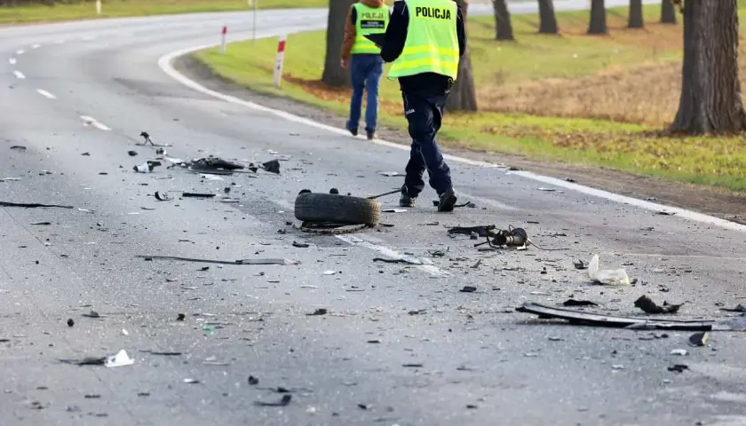 Na drodze po wypadku leżą szczątki pojazdów i opona. Policjanci w kamizelkach odblaskowych badają miejsce zdarzenia, być może szukając śladów ucieczki z miejsca wypadku.