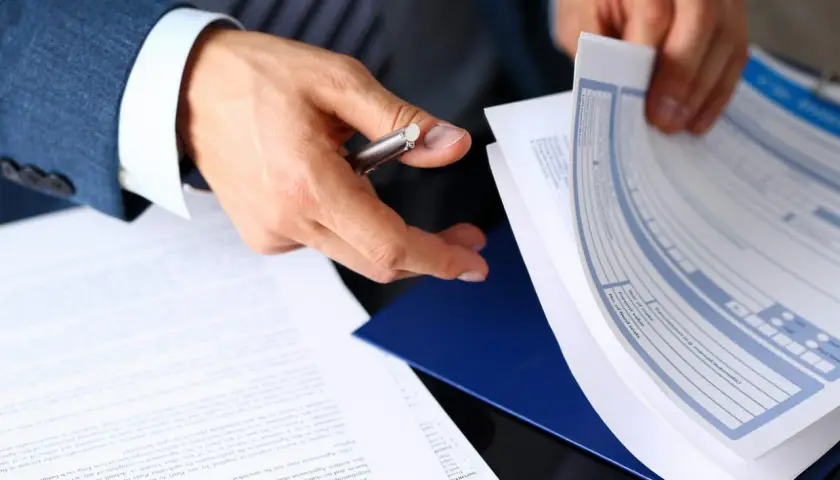 Ręce mężczyzny w garniturze przeglądają dokumenty, wśród których znajduje się formularz ubezpieczenia pośrednika nieruchomości.