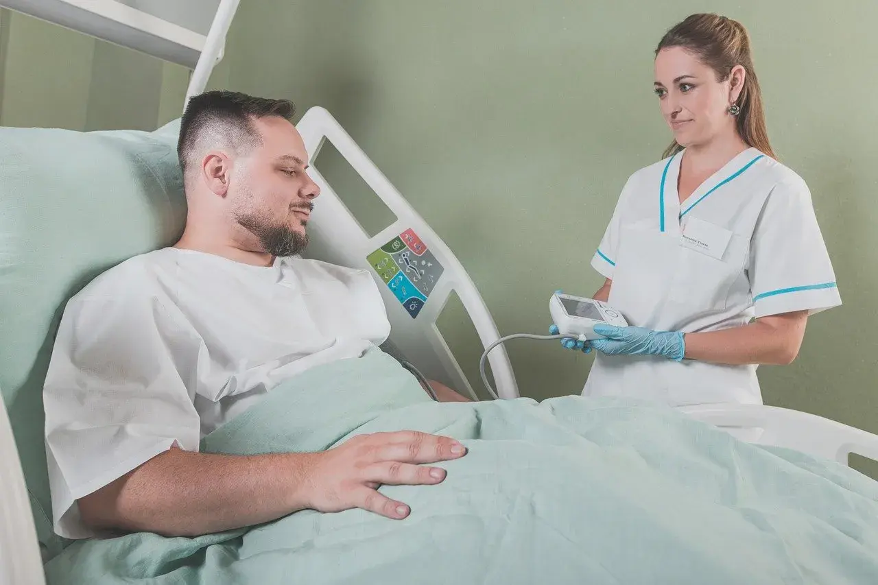 Uśmiechnięta pielęgniarka z aparatem do pomiaru ciśnienia w rękach, pacjent w łóżku szpitalnym. Radość z zakończenia leczenia.