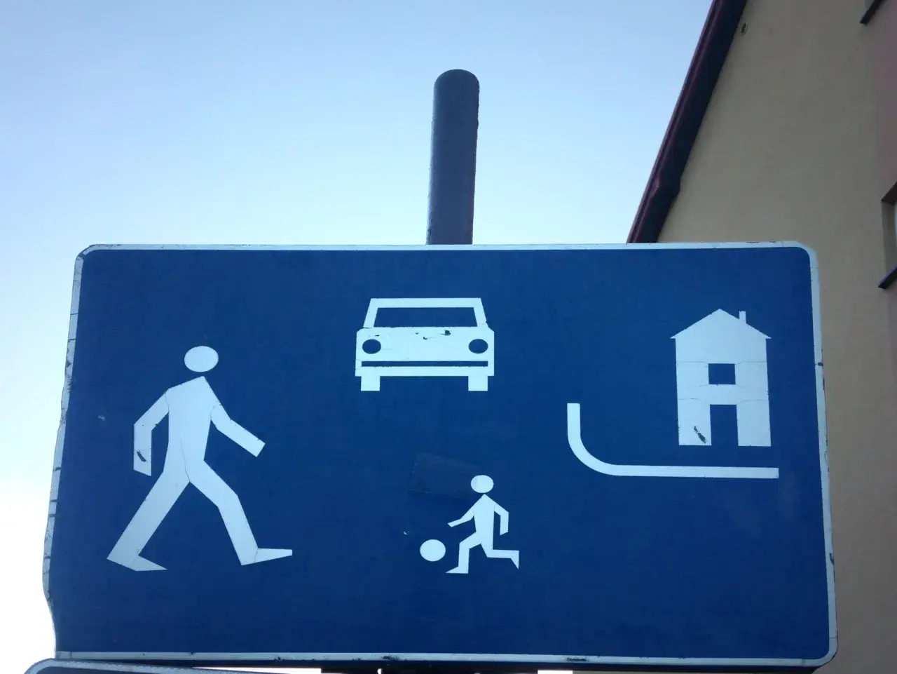 Znak informuje, że w strefie ruchu pieszy ma pierwszeństwo. Widać pieszego, dziecko grające w piłkę, samochód i dom.