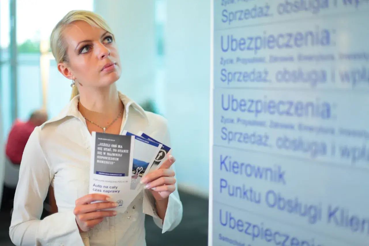 Kobieta z ulotkami zastanawia się nad przedłużeniem OC przed terminem. Na tablicy widnieje napis "Ubezpieczenia".