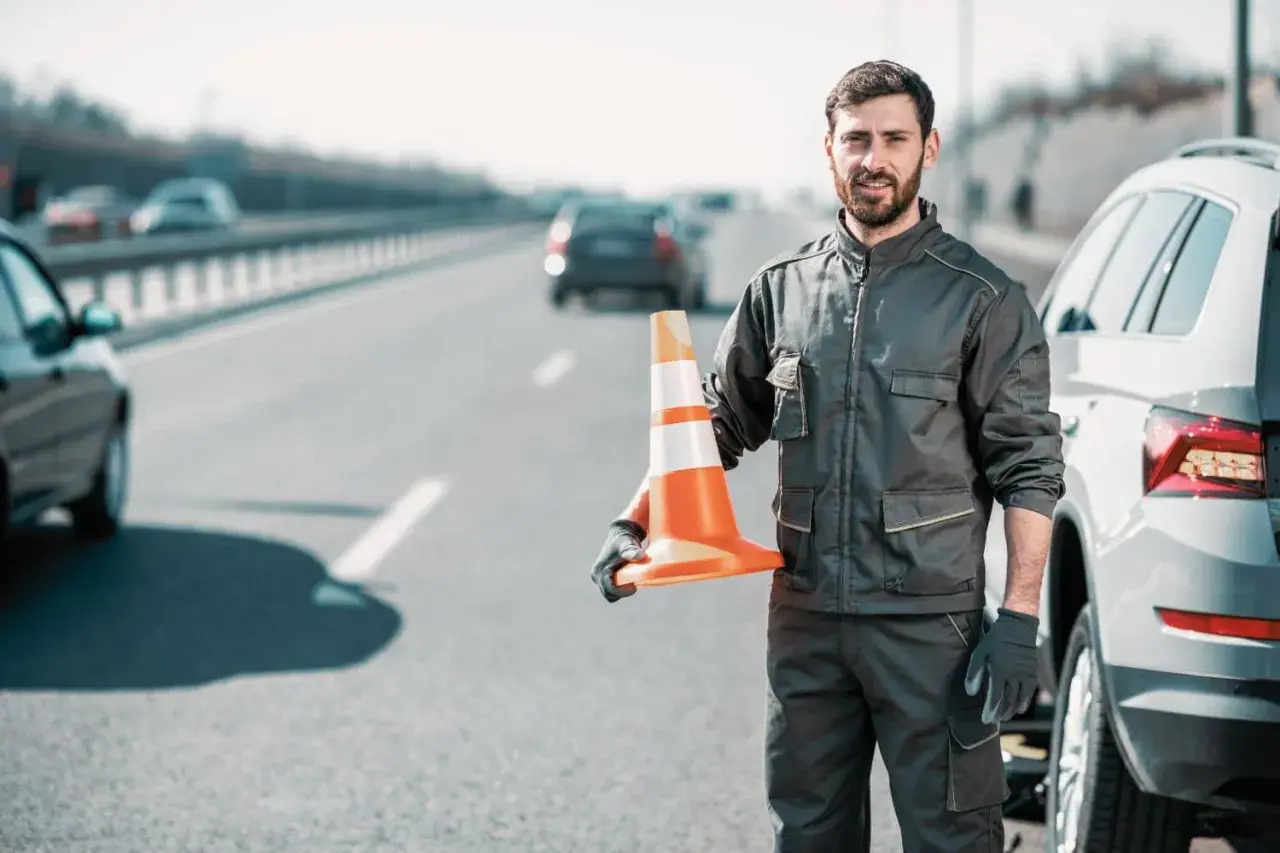 Pomoc drogowa na autostradzie. Mężczyzna w roboczym ubraniu trzyma pomarańczowy pachołek, gotowy do zabezpieczenia miejsca awarii auta.