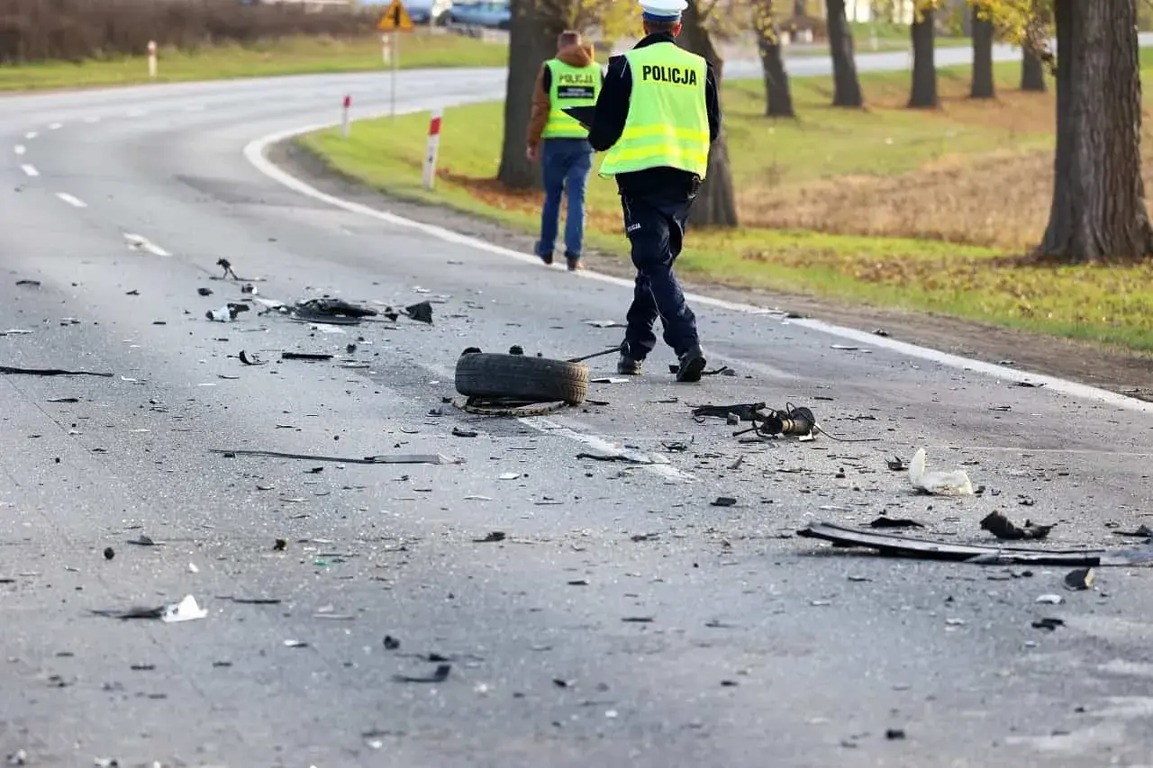 Na drodze po wypadku leżą szczątki pojazdów i opona. Policjanci w kamizelkach odblaskowych badają miejsce zdarzenia, być może szukając śladów ucieczki z miejsca wypadku.
