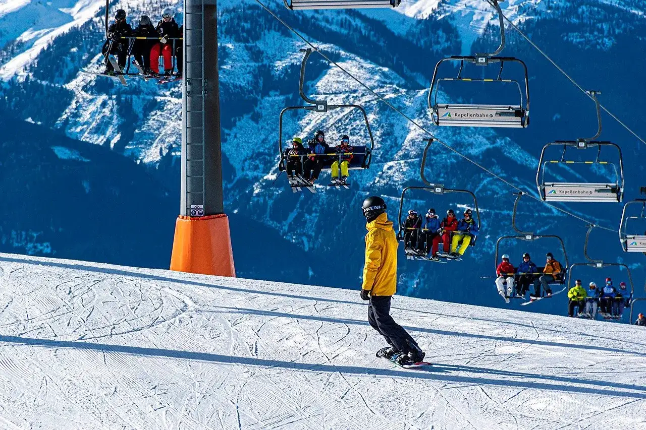Snowboardzista w ż&oacute;łtej kurtce zjeżdża po stoku, podczas gdy na wyciągu krzesełkowym siedzą narciarze. W tle g&oacute;ry.
