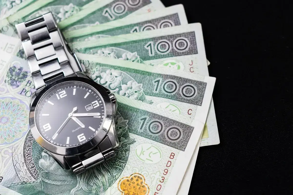 Zegarek na stosie banknotów 100 zł. Czas to pieniądz, zwłaszcza gdy chodzi o opłacanie składek ZUS samemu.