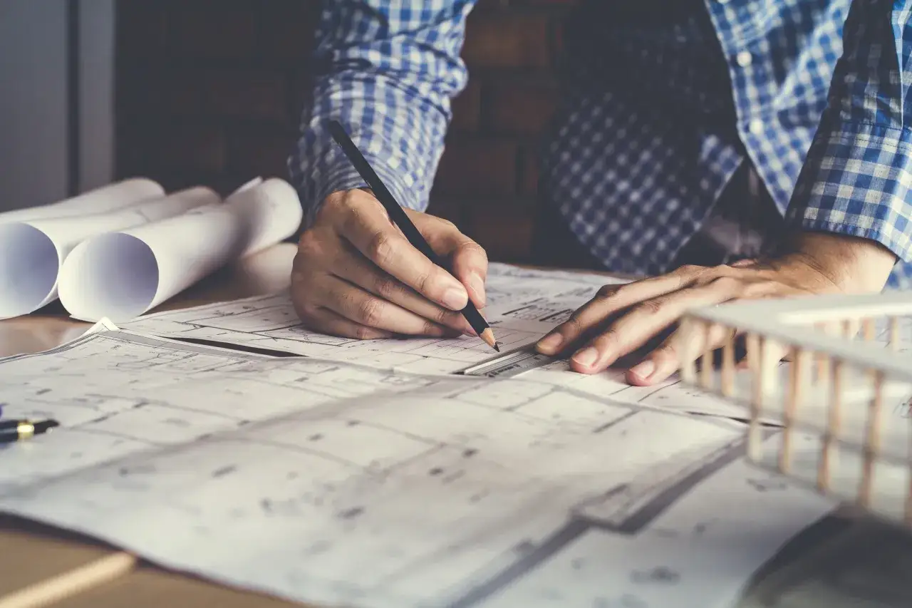 Ręce projektanta pracują nad szkicami, tworząc wizję przyszłego budynku. Obok leżą zwinięte plany i makieta.