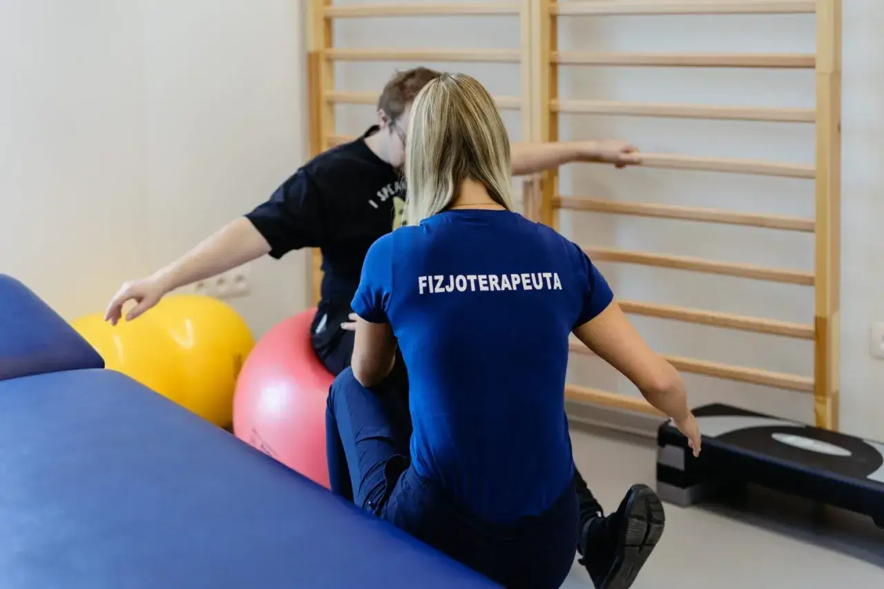 Fizjoterapeutka pomaga pacjentowi po złamaniu nadkłykciowym kości ramiennej w rehabilitacji. Ćwiczenia z piłkami i drabinką.