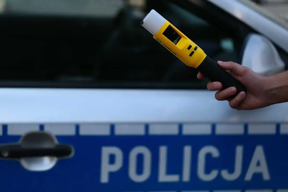 Ręka trzyma żółty alkomat na tle radiowozu z napisem POLICJA. To rutynowa kontrola trzeźwości.