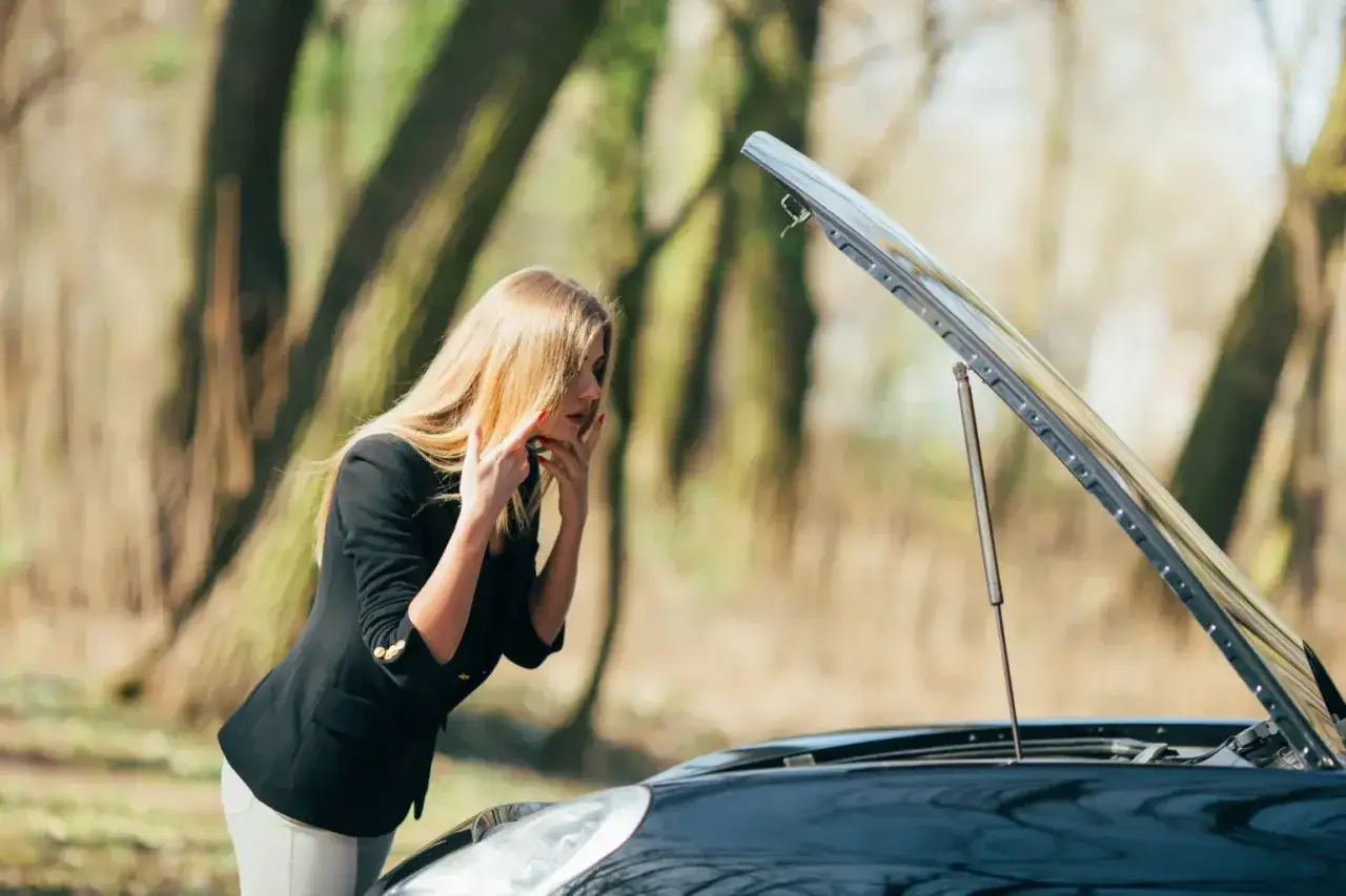 Kobieta z blond włosami patrzy na otwartą maskę samochodu, zmartwiona. Awaria auta na autostradzie.