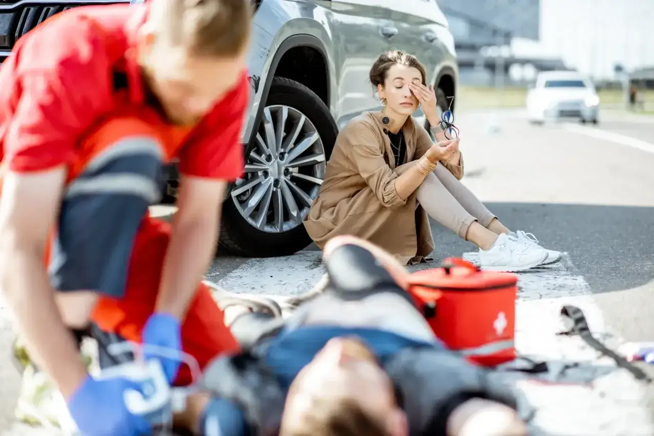 Po wypadku drogowym, ratownik udziela pomocy poszkodowanemu, podczas gdy kobieta siedzi obok, wstrząśnięta. Wypadki dzielimy na te, które wymagają natychmiastowej interwencji.