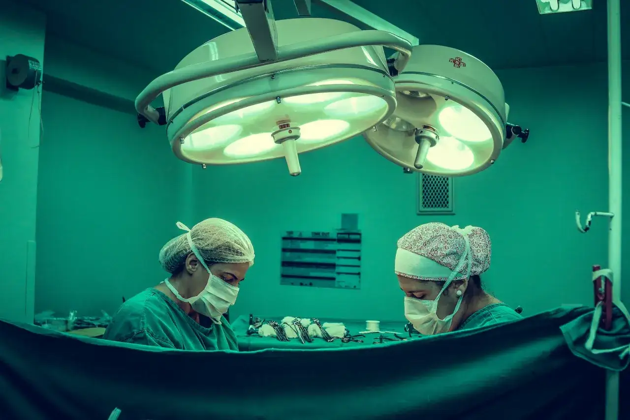 Dwie lekarki w maseczkach i czepkach pracują pod lampami operacyjnymi. Czekają na odszkodowanie za operacje.