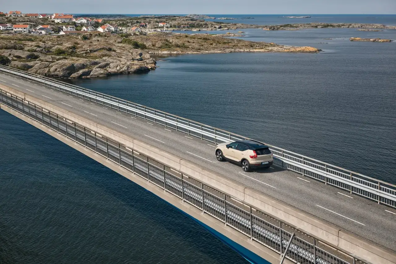 Beżowy SUV jedzie mostem nad wodą w kierunku norweskiej wioski. Pomyśl o ubezpieczeniu w Norwegii, podr&oacute;żując po malowniczych drogach.