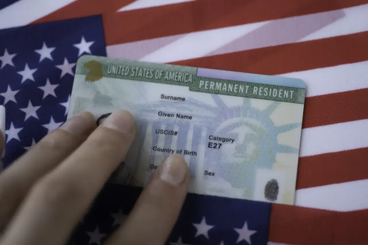 Dłoń trzyma zieloną kartę, dokument potwierdzający status stałego rezydenta USA, na tle amerykańskiej flagi. To dow&oacute;d, co to zielona karta.
