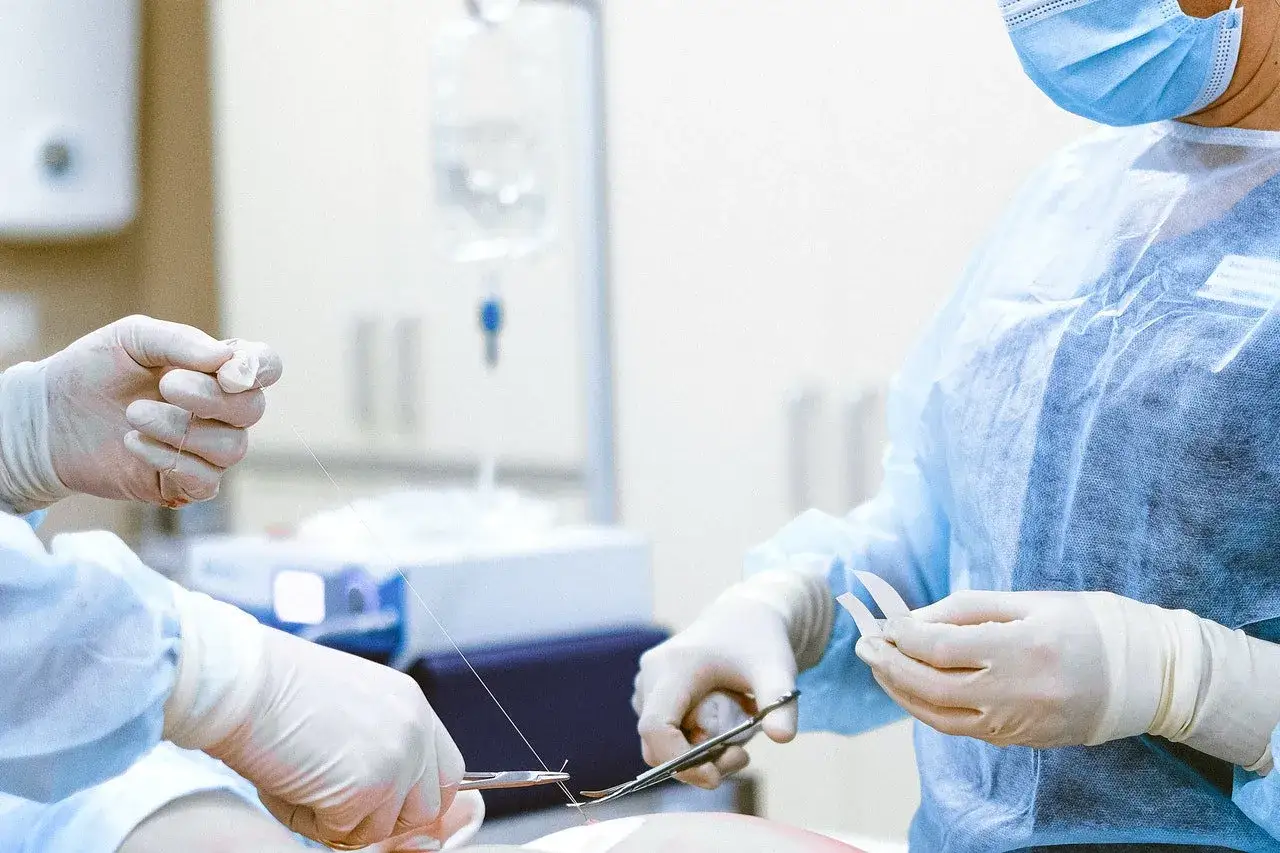 Chirurgiczne ręce w rękawiczkach szyją ranę. W tle kroplówka, ważna dla pacjenta, który może ubiegać się o odszkodowanie za operację.