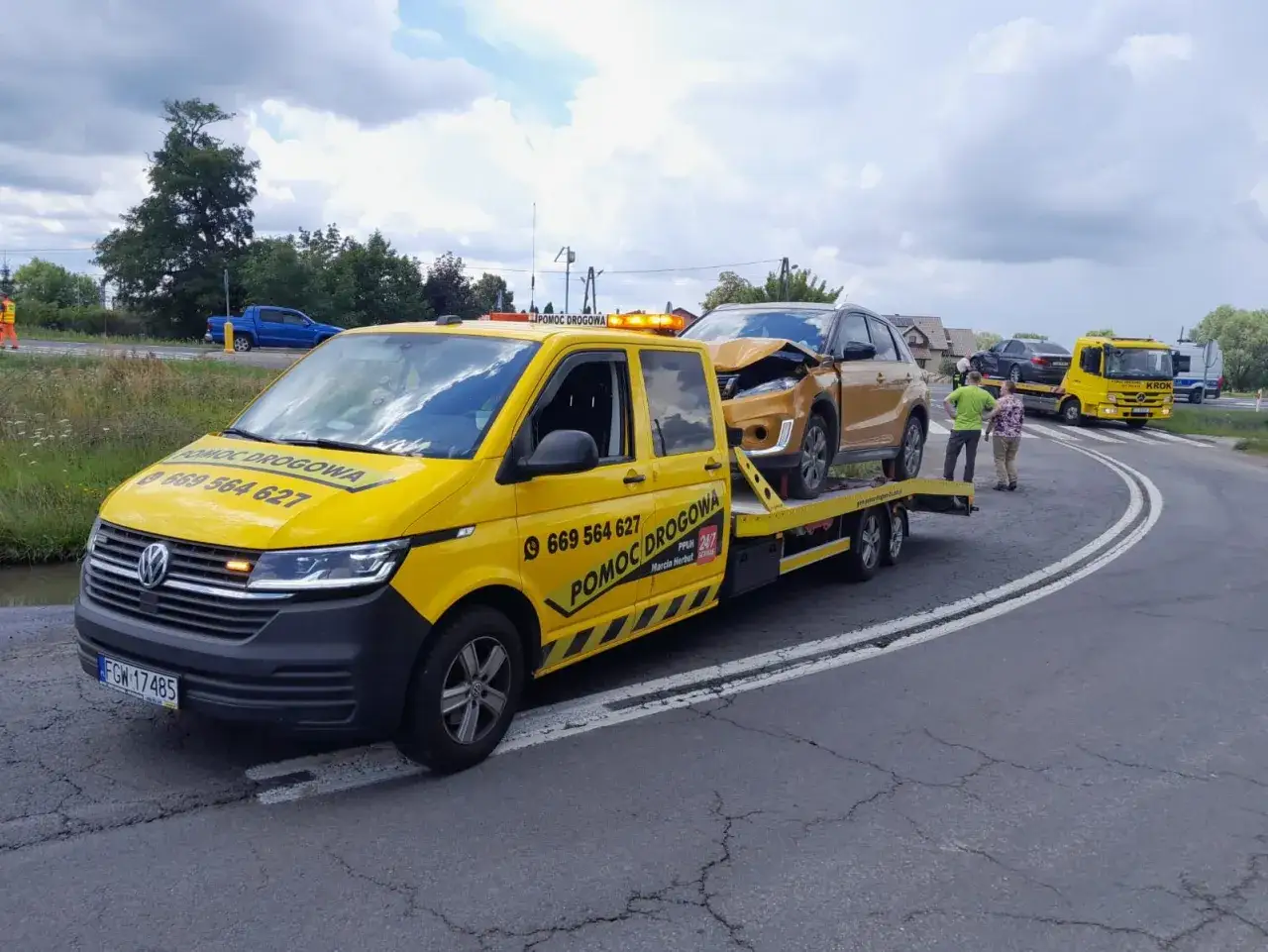 Żółty pojazd pomocy drogowej z uszkodzonym złotym autem na lawecie. W tle inne pojazdy i ludzie. Usługa mtu assistance w akcji.